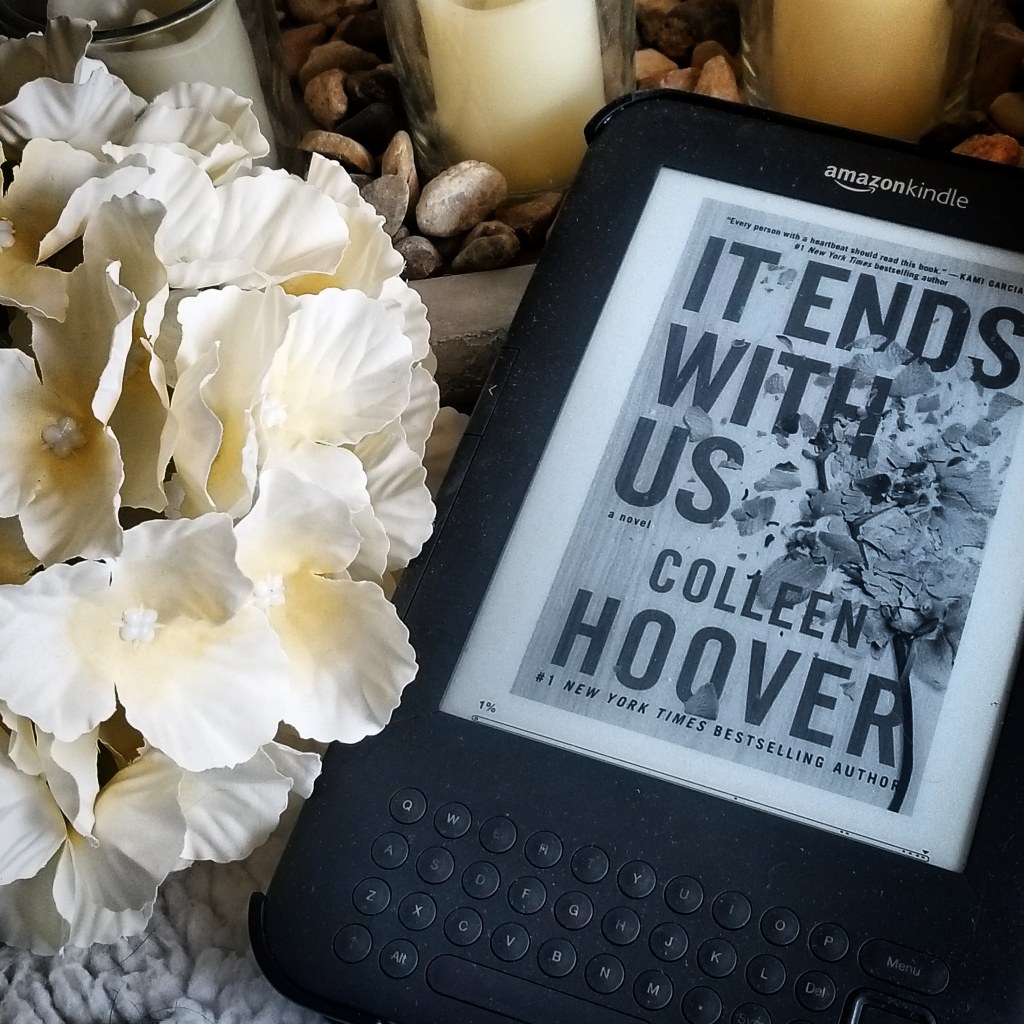 Photo of a Kindle keyboard ereader displaying It Ends With Us by Colleen Hoover next to a bouquet of white flowers and below three small candles.