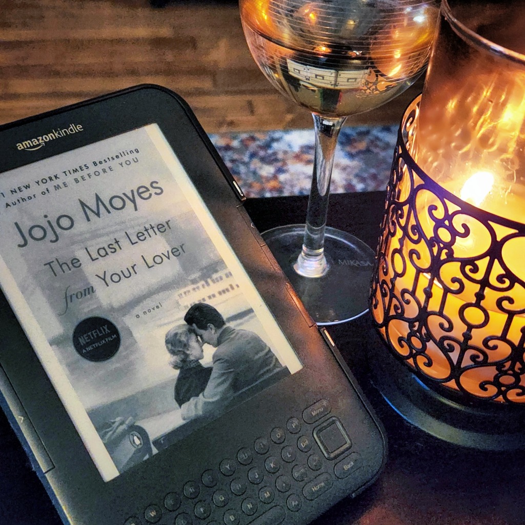 A Kindle ereader showing the ebook cover of The Last Letter from Your Lover by Jojo Moyes on a coffee table next to a glass of white wine and a burning candle.