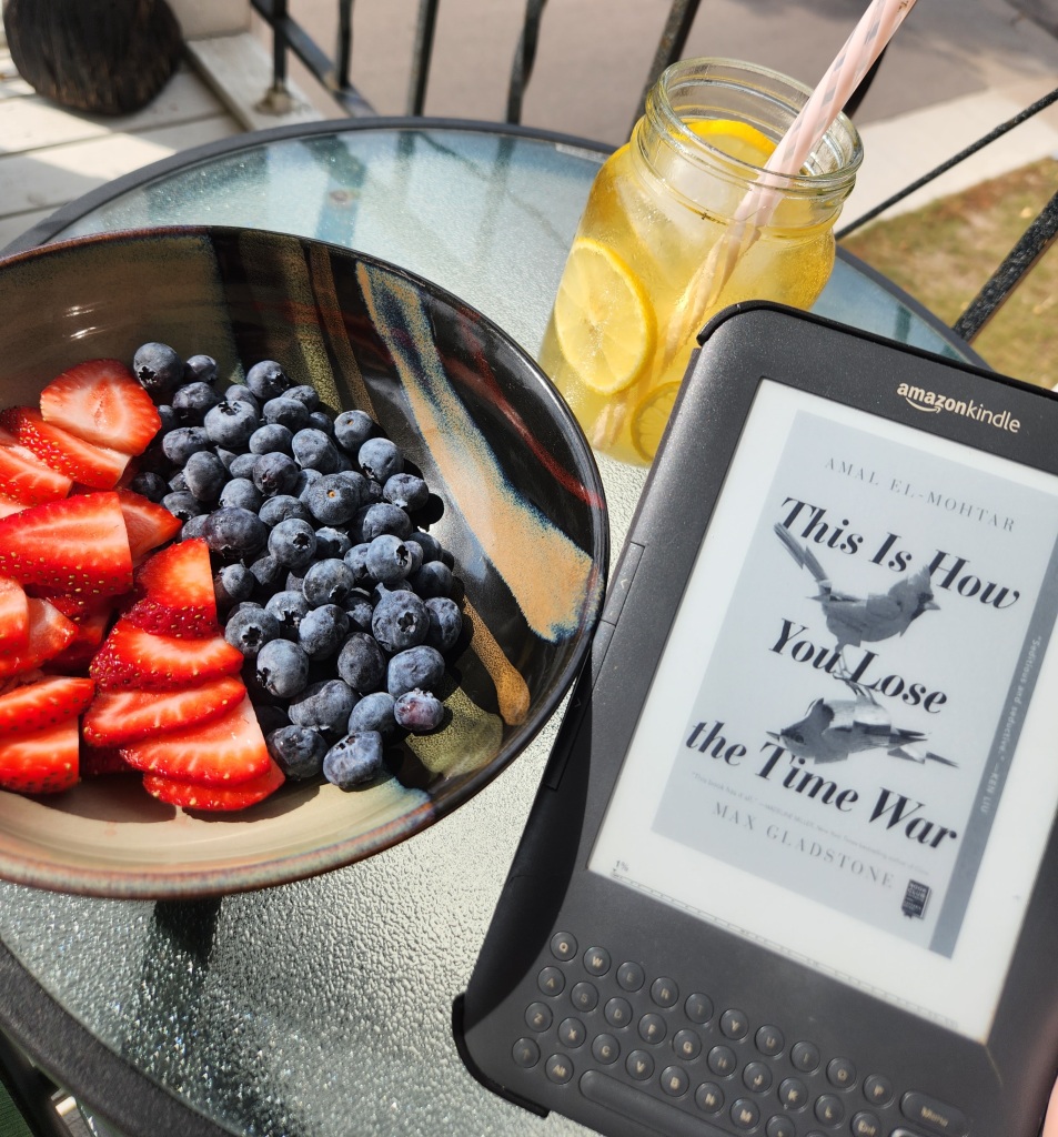 A Kindle keyboard with the cover of This Is How You Lose the Time War by Amal El-Mohtar and Max Gladstone on a round table with a bowl of cup up strawberries and blueberries and a mason jar of iced tea and lemon slices with a straw in the sunshine.